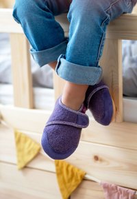 Paarse instapschoenen met een zachte fleecetextuur en verstelbare band, gecombineerd met opgerolde denim jeans, rustend op een houten bedframe.