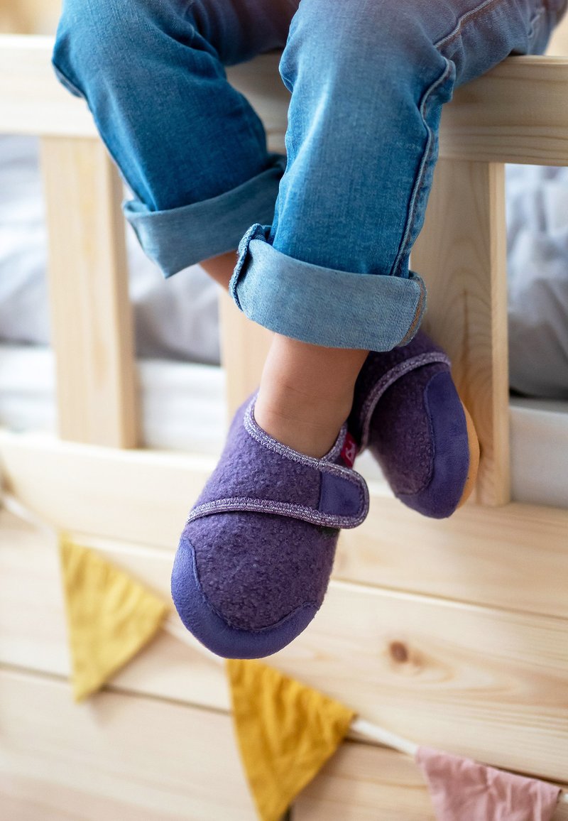 Paarse instapschoenen met een zachte fleecetextuur en verstelbare band, gecombineerd met opgerolde denim jeans, rustend op een houten bedframe.