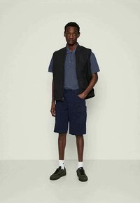 Marineblå poloshirt, svart vest med glidelås og marineblå cargoshorts. Sorte joggesko og hvite sokker fullfører antrekket. Glatte teksturer og avslappet design.