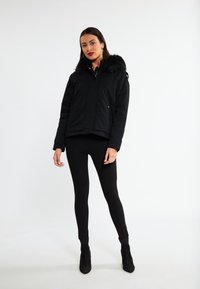 Sort polstret jakke med kunstpelskapuze, med lynlås foran og gyldne detaljer. Bæret over sorte leggings og sorte ankle boots med hæl.