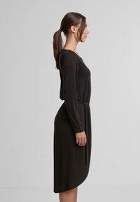 Schwarz langärmliges Kleid mit runder Ausschnitt, elastischem Taillenbund und asymmetrischem Saum. Der Stoff hat eine sanfte Textur.