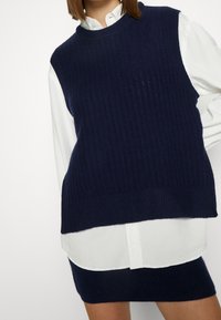 Jersey sin mangas de color azul marino con textura de canalé y cuello redondo, superpuesto sobre una camisa blanca con cuello, mostrando colores contrastantes.