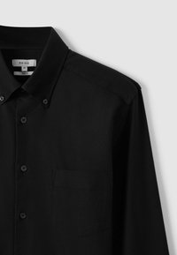 Cămașă neagră slim-fit fabricată dintr-un material neted. Are guler cu nasturi, nasturi frontali și un buzunar pe piept.