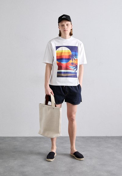 White T-shirt with multicolored sunset graphic, dark denim shorts, black canvas slip-ons, and beige tote bag with brown handles.