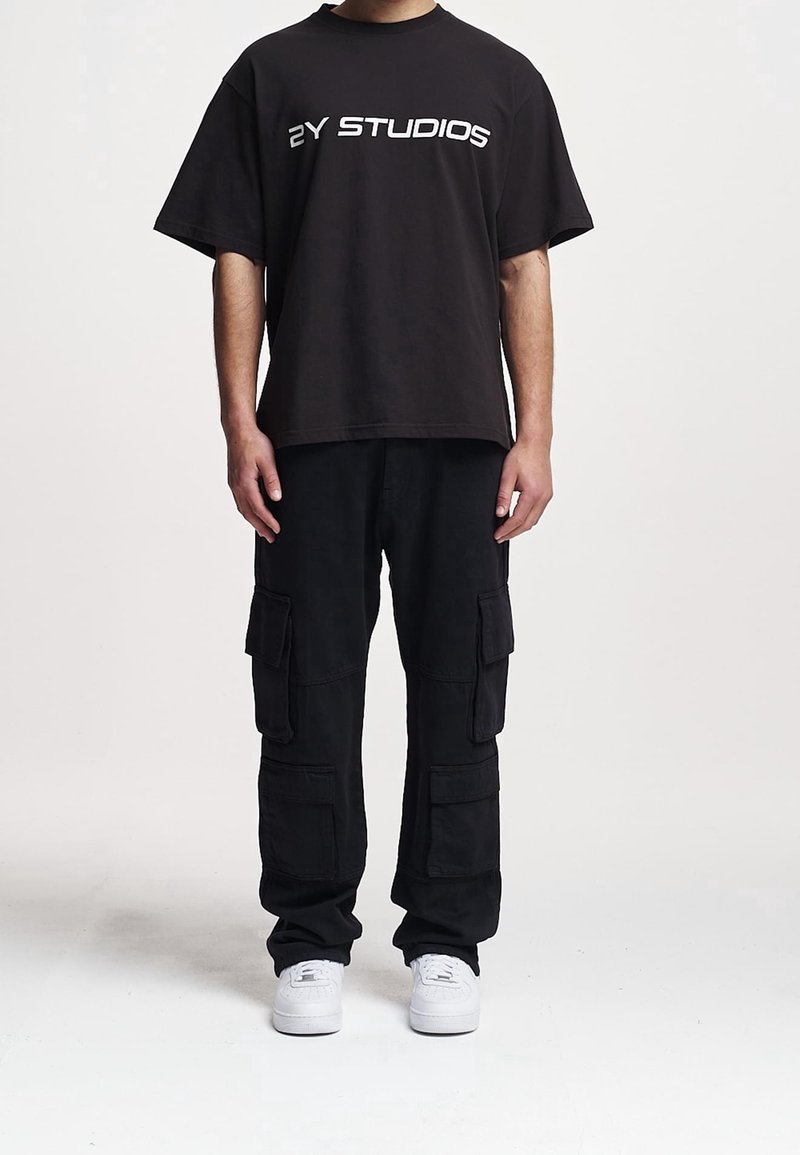 T-shirt noir oversize avec l'inscription "ZY STUDIOS" en blanc. Associé à un pantalon cargo noir à multiples poches. Des baskets blanches apportent un contraste.