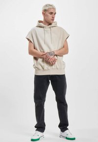 Beige ärmelloser Hoodie mit Kordelzug und vorderer Tasche, kombiniert mit dunkelblauen Jeans und weißen Sneakern mit grünen Akzenten.