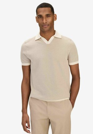 Man som står med ena handen i fickan, klädd i en beige kortärmad stickad pikétröja och matchande ljusbeige byxor mot en vit bakgrund.