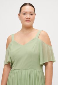 Lichtgroen jurk met een V-hals en off-shoulder fladdermouwen. Gemaakt van een vloeiende stof met geplooide textuur rond de taille.