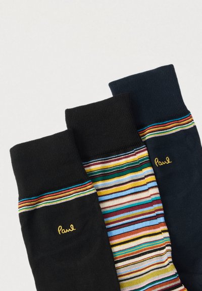 Trois paires de chaussettes : une noire unie, une bleu marine foncé et une avec des rayures multicolores. Chaque paire a une bordure côtelée et porte l'inscription "Paul" brodée en jaune.