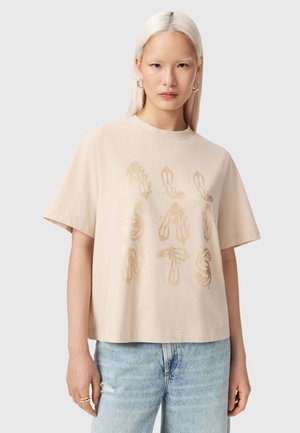 Kvinde med glat platinblondt hår iført oversized beige T-shirt med guldfarvede skooptryk og lyseblå slidt jeans, neutral baggrund.