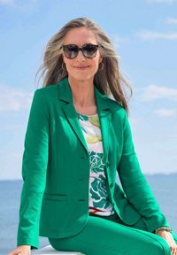 Grüner maßgeschneiderter Blazer und passende Hose über einem floral gemusterten Hemd, accessoriert mit runden Sonnenbrillen und Creolen. Ozeanhintergrund.