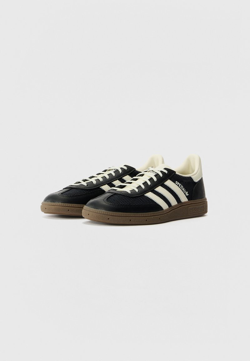 Baskets de sport noires avec des rayures blanches, tige en tissu texturé, semelle en caoutchouc et accents beiges contrastants au niveau du talon et de la languette.