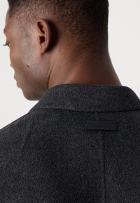 Donkergrijze wollen jas met een strakke uitstraling, met notch-kraag en een zwart merklabel op de achterkant. De textuur lijkt iets pluizig.