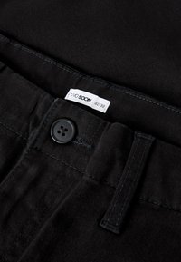 Svarte denimbukser med glatt tekstur, med en lukking med fire knapper, kontrasterende sømmer, og en etikett som angir størrelse 32/32.