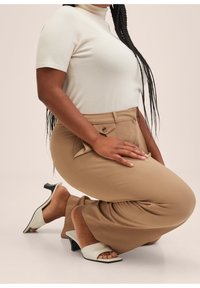 Pantalons beiges bien taillés avec poches latérales, col roulé ajusté crème et sandales à talons ouverts avec une bande blanche et un talon noir.