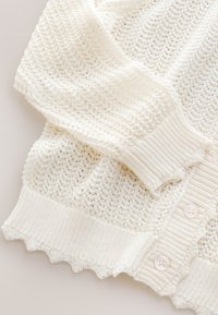 Kremo spalvos megztinis su sagomis, ribotu rankogaliu ir banguotu apdailos kraštu, pasižymintis oriu atviru raištelių raštu.