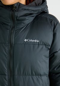 Černá péřová bunda s kapucí, s předním zipem a logem Columbia. Materiál vypadá hladce a lehce s prošívaným vzorem.