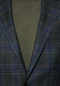 Dwie warstwy odzieży: ciemnozielona bluzka z okrągłym dekoltem oraz ciemny blazer w niebiesko-brązową kratę, z teksturowanym materiałem i klapami.