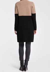 Strikket cardigan i beige, sort og tan farveblokdesign. Lange ærmer, midt på låret længde, tekstureret materiale. Båret over mørke jeans og støvler.