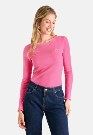 Frau lächelt und schaut nach unten, trägt ein pinkes langärmeliges Oberteil und dunkelblaue Jeans vor einem schlichten hellen Hintergrund.