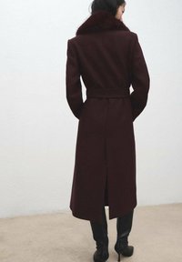 Bordeauxrode wollen jas met een kraag van nepbont, een ceintuur in de taille en een detail van een achterventilatie, met een getailleerde pasvorm en lange mouwen.