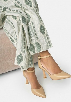 Jambes croisées portant un pantalon blanc brodé de feuilles vertes et des escarpins beige à bout pointu avec bride à la cheville, assise sur une chaise rose clair.