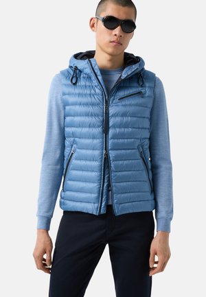 Helleblaue Pufferweste mit figurschmeichelndem Design, Fronttaschen mit Reißverschluss und Kordelzugkapuze. Getragen über einem langärmligen blauen Shirt.