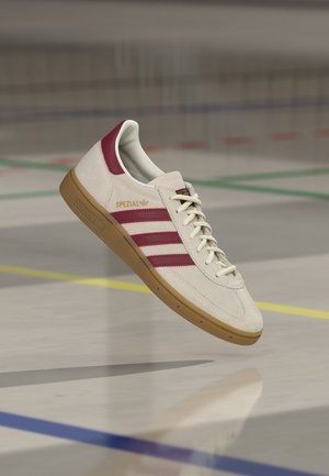 Zapatilla blanca adidas Spezial con rayas granate y pestaña en el talón, suela de goma, con cordones, flotando sobre un suelo de gimnasio con líneas de colores.