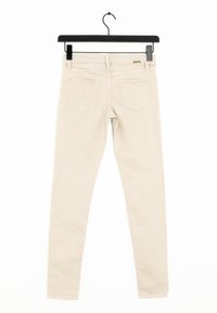 Beige skinny jeans hangen aan een zwarte hanger, met een gladde textuur, vijf zakken en een subtiele logo-detail op de tailleband.