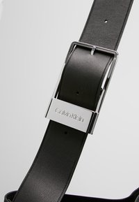 Czarny skórzany pasek z polerowaną srebrną klamrą, na której wygrawerowano logo "Calvin Klein". Pasek ma gładką fakturę i precyzyjnie wykonane dziurki.