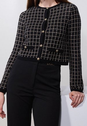 Femme portant un cardigan à carreaux noir et blanc avec des boutons dorés et un pantalon noir taille haute, posant avec une main posée sur une surface.