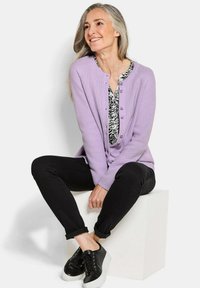 Vrouw met lang grijs haar die glimlacht, gekleed in een lila cardigan over een gemusterde blouse, zwarte broek en zwarte sneakers, zittend op een wit blok.