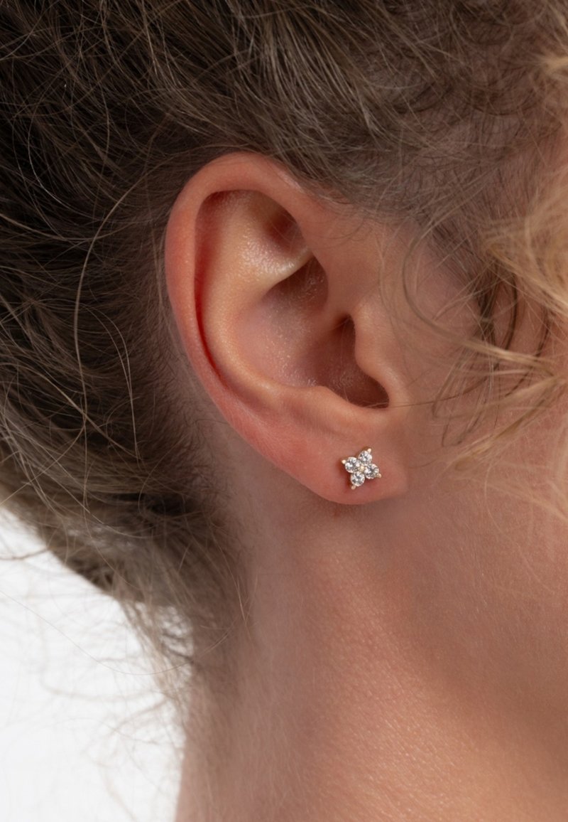 Zilveren studoorbel in bloemblaadvorm, versierd met heldere rhinestones. Tegen een neutrale huidtint en een achtergrond van golvend haar.