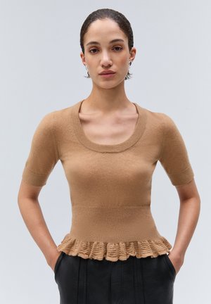 Femme aux cheveux courts et foncés porte un top en maille beige à manches courtes avec un ourlet volanté et un pantalon noir, debout devant un fond clair uni.
