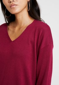 Vinröd v-ringad tröja i ett mjukt stickat tyg med avslappnad passform och ribbade detaljer vid halslinningen. Stora silvriga hoopsörhängen syns.