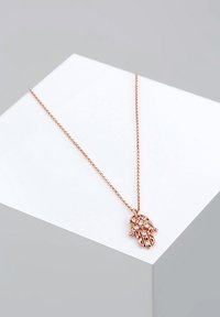 Halskette aus roségold mit einem handförmigen Anhänger, verziert mit klaren Kristallen, aufgehängt an einer zarten Kette. Die flache Oberfläche darunter hebt das Design hervor.