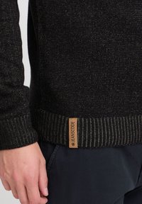Zwarte gebreide trui met gestructureerde stof, geribbelde zoom en een contrasterend leren label met "JEANSCODE." Close-up van de mouw.