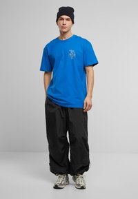 Blå bomulds T-shirt med et grafisk print på brystet. Kombinationen med sorte, løstsiddende bukser og grå træningssko.