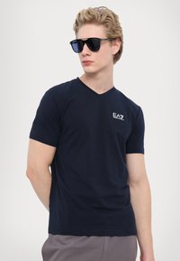 Námořnické tričko s V-výstřihem vyrobené z měkké látky, krátké rukávy, s malým bílým logem na hrudi. Model má také na sobě tmavé sluneční brýle.