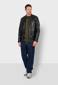 Černá kožená bunda s vysokým límečkem, prošívané ramenní akcenty, nošená přes olivově zelený top a modré džíny, doplněná o světlé tenisky.