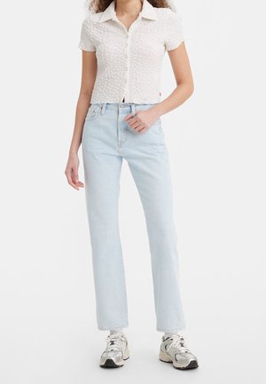 Femme portant une chemise blanche texturée à manches courtes, un jean droit taille haute bleu clair et des baskets blanches, debout devant un fond uni.