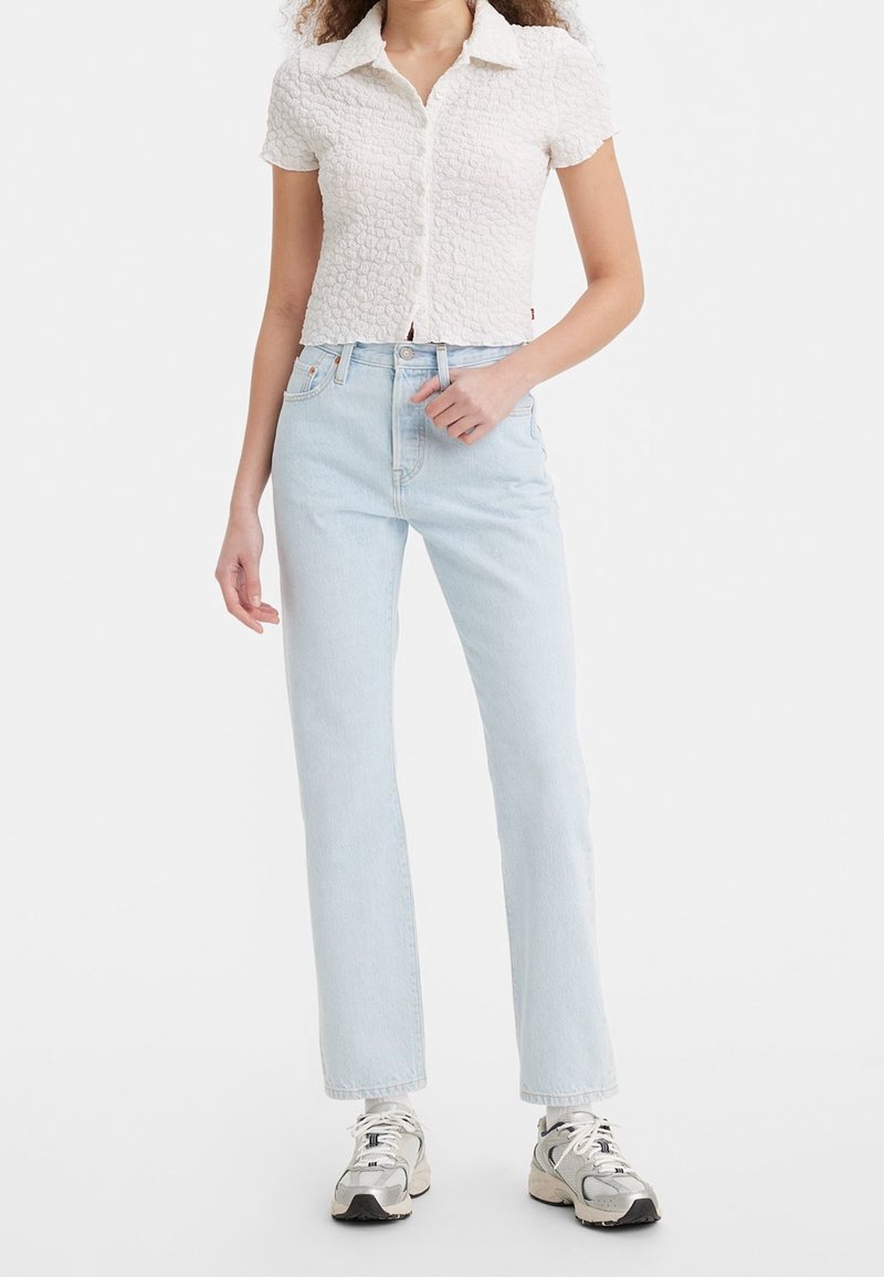 Femme portant une chemise blanche texturée à manches courtes, un jean droit taille haute bleu clair et des baskets blanches, debout devant un fond uni.