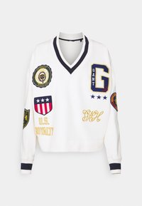 Sweat-shirt blanc avec accents bleu marine, col en V, et plusieurs patchs brodés en vert, rouge et or, représentant des logos et des détails.