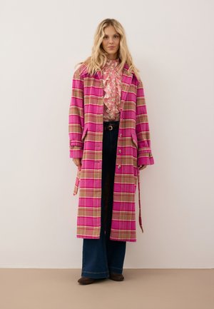Lang roestkleurige geruite jas met tan accentuering, voorzien van knoopsluitingen, zijzakken en een tailleriem. Onder de jas is een bloemenblouse zichtbaar.