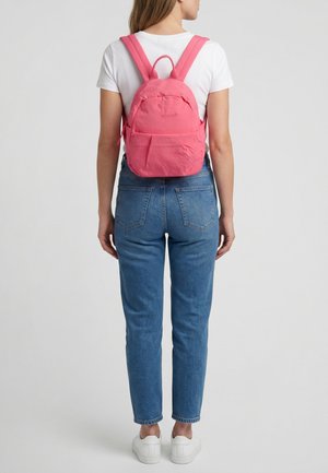 Vrouw draagt wit T-shirt, blauwe jeans, witte sneakers en een kleine roze rugzak, staat met haar rug naar ons toe op een effen achtergrond.