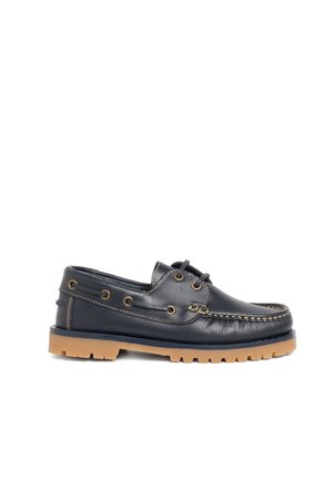Zapato de barco de cuero negro con suela de goma marrón y cordones negros a juego pasados por ojales de latón, vista lateral sobre fondo blanco.