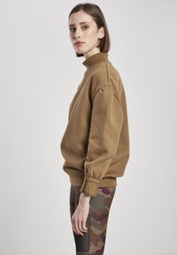 Sweatshirt maro cu gât înalt, mâneci voluminoase și manșete cu nervuri. Purtați cu colanți camuflați în nuanțe de maro și verde.