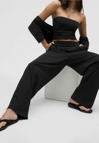 Femme portant un pantalon noir à jambes larges, un crop top bustier et des tongs, assise jambes écartées sur un fond blanc.
