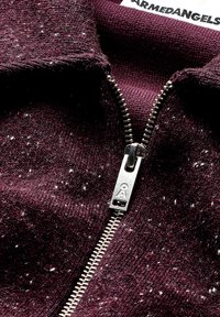 Burgunderfarbene Zip-Jacke mit einem metallischen Reißverschluss, strukturiertem Strickstoff mit Einsprengseln und gerippten Bündchen. Markenetikett an der Kragen sichtbar.
