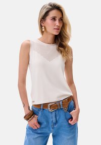 Witte mouwloze top met een gaasdetail in een V-vorm aan de voorkant. Gecombineerd met lichtblauwe jeans en een bruine riem met een decoratieve gesp.